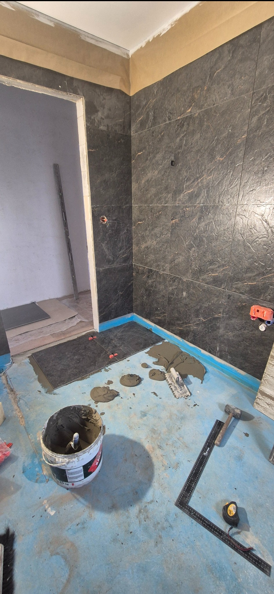 Układanie ciemnych płytek imitujących kamień na ścianach i podłodze łazienki. Widoczne narzędzia glazurnicze i wiadro z zaprawą. Niebieska folia w płynie na podłodze.