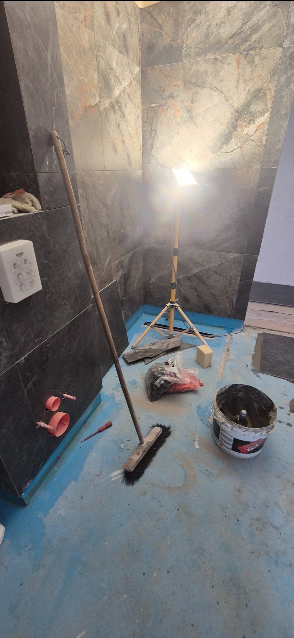 Etap prac w łazience: ciemne płytki na ścianach, niebieska podłoga, widoczne narzędzia (szczotka, wiadro z zaprawą), oświetlenie budowlane. Przygotowanie do dalszych prac wykończeniowych.
