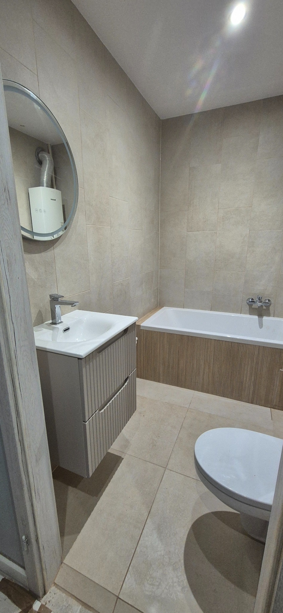 Jasna łazienka po remoncie: beżowe płytki, umywalka z szafką o ryflowanych frontach, wanna obudowana drewnopodobną płytką, okrągłe lustro z widocznym piecykiem.