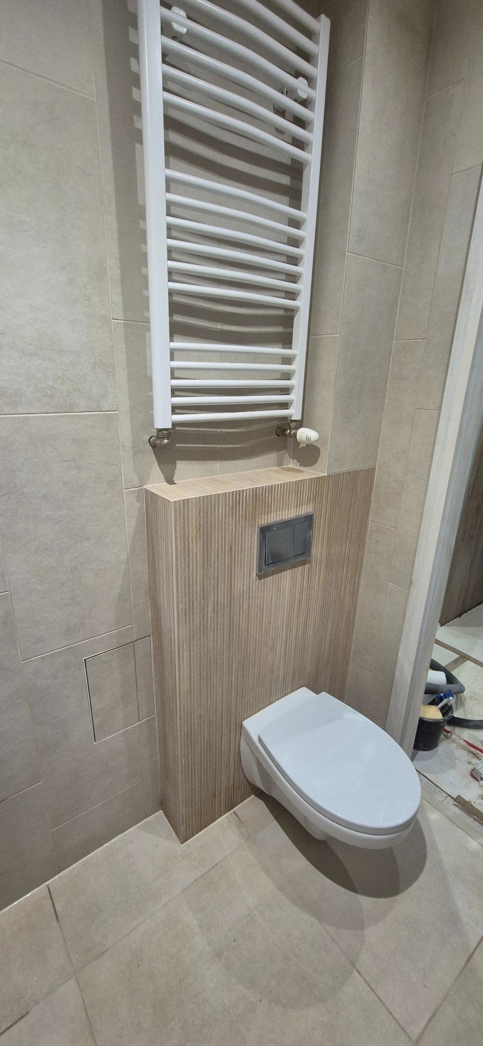 Nowoczesna łazienka po remoncie: beżowe płytki, podwieszana toaleta z drewnianą obudową i biały grzejnik drabinkowy. Widoczne narzędzia sugerują trwające prace wykończeniowe.