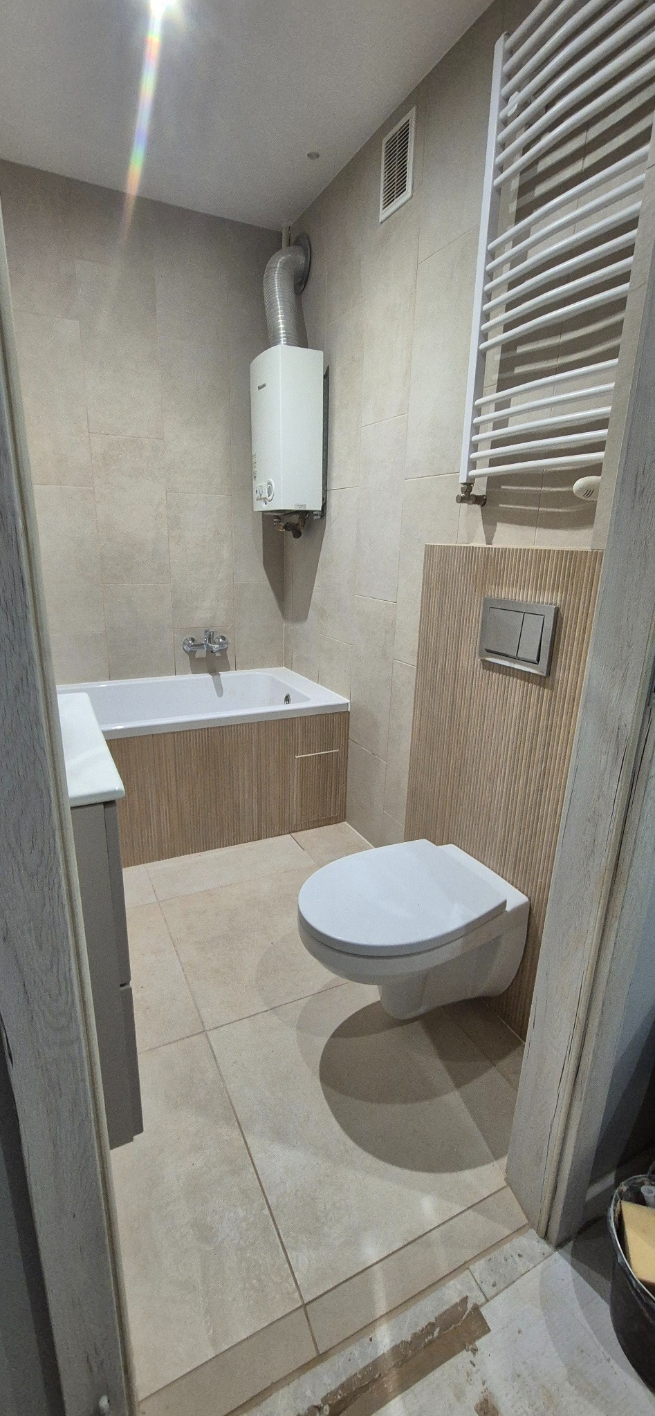 Jasna łazienka po remoncie: beżowe płytki, podwieszana toaleta z panelem, grzejnik drabinkowy, wanna z obudową i piecyk gazowy. Widok zza futryny.