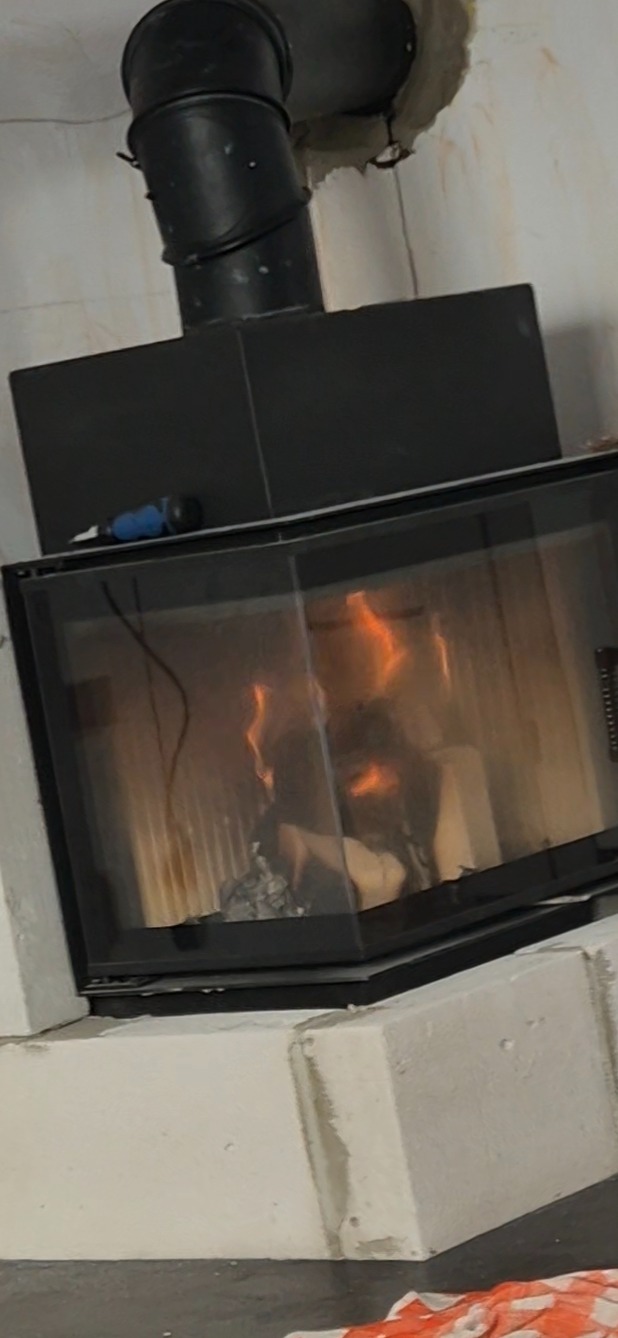 Opalający się kominek z czarnym wkładem i widocznym przewodem kominowym. Na górze kominka leży niebieski śrubokręt. Widoczne białe bloczki fundamentowe.