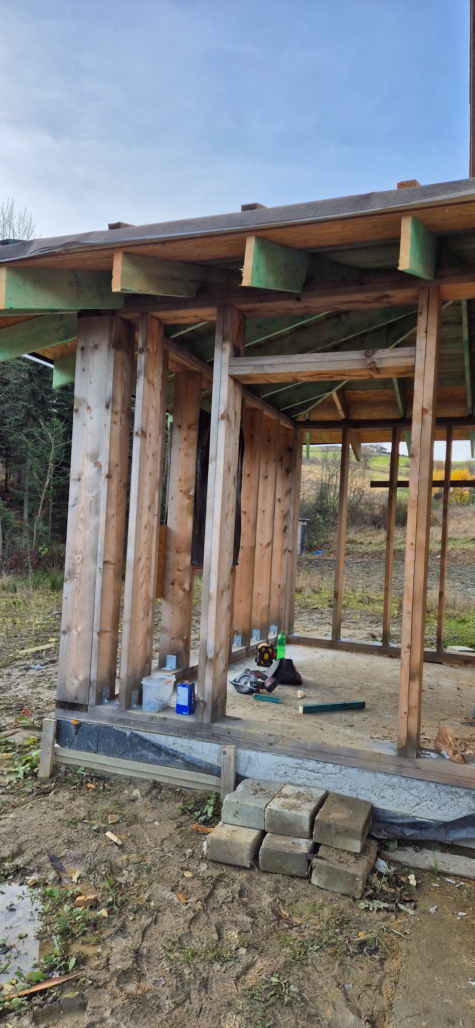 Surowa konstrukcja drewniana z widocznymi narzędziami i materiałami budowlanymi na placu budowy. Widoczne elementy dachu i ścian w trakcie wznoszenia.