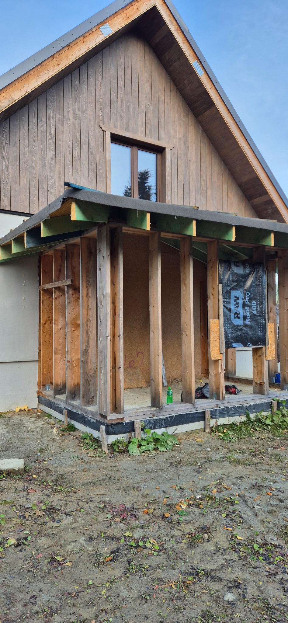 Budowa drewnianego domu w trakcie realizacji. Widoczne drewniane słupy i konstrukcja dachu. Częściowo ocieplona ściana. Na zewnątrz ziemia i roślinność.