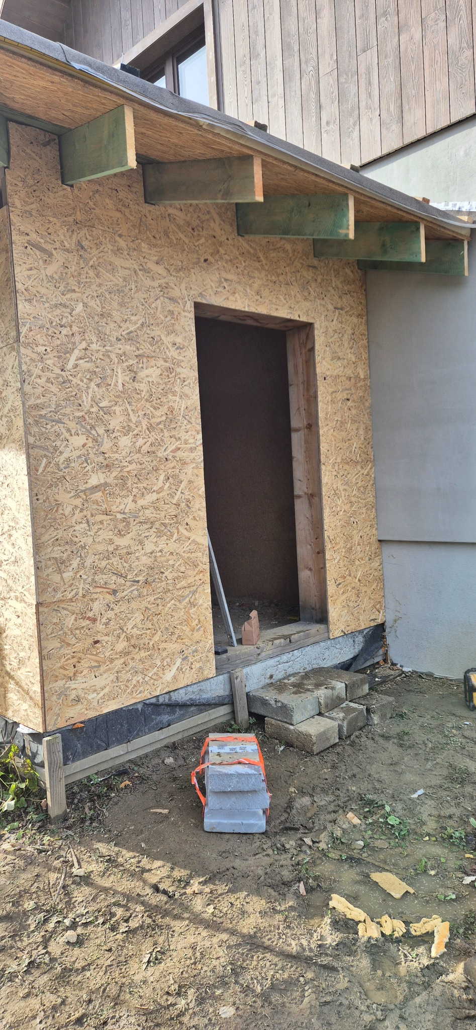 Budowa ganku z płyt OSB w Żorach. Widoczne surowe drewno, bloczki betonowe i ziemia. Etap prac przed wykończeniem elewacji.