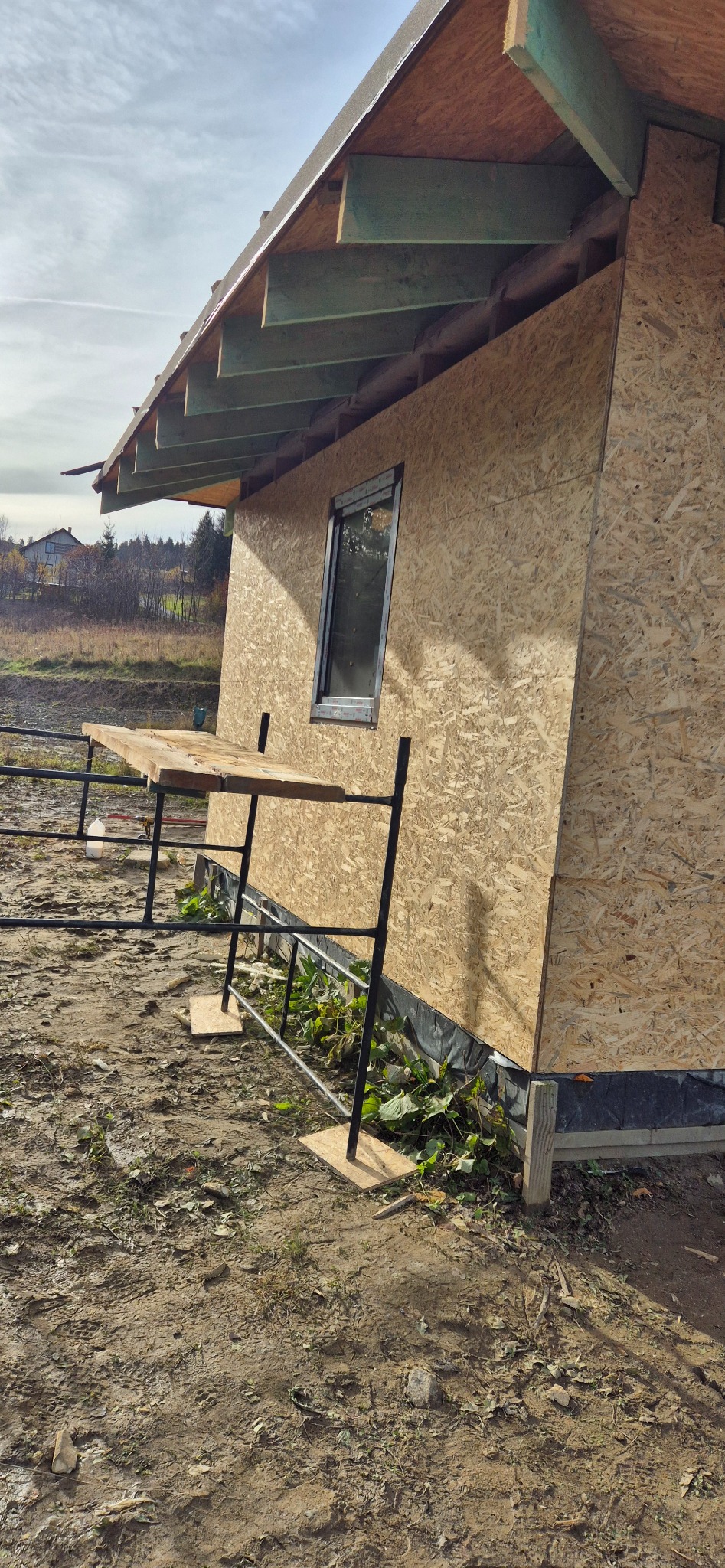 Budowa szkieletowa domu z płyt OSB, widoczne okno i rusztowanie. Prace w toku, teren wokół niezagospodarowany. Ujęcie z dołu, podkreślające konstrukcję dachu.