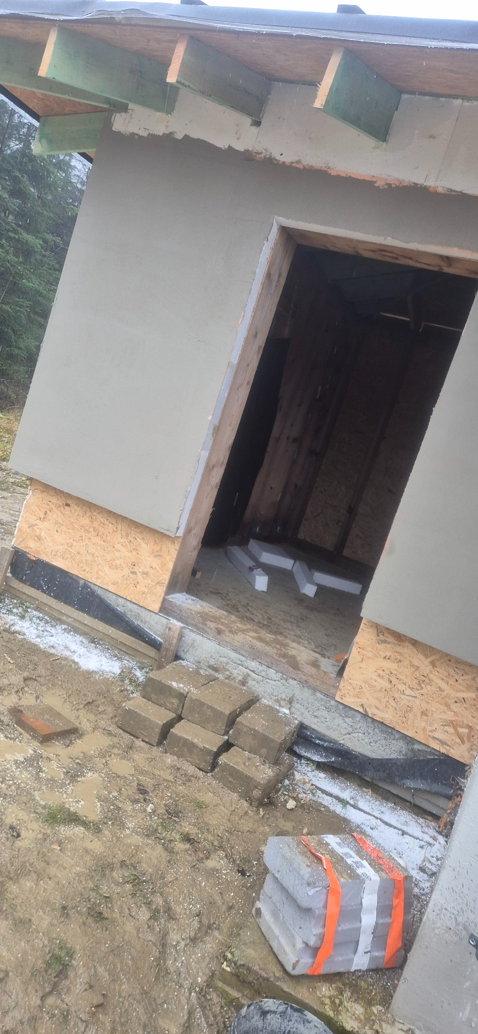 Surowy stan elewacji budynku z oknem bez ramy, widoczne materiały budowlane i błoto. Etap prac wykończeniowych w trakcie realizacji.