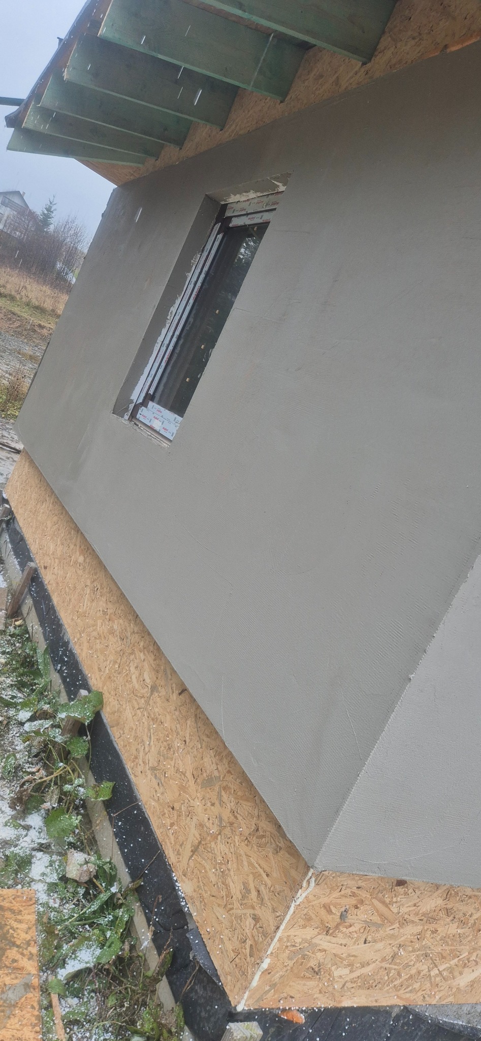 Świeżo otynkowana ściana zewnętrzna budynku z zamontowanym oknem, widoczne deski szalunkowe i fragment dachu. Szara elewacja w surowym otoczeniu.