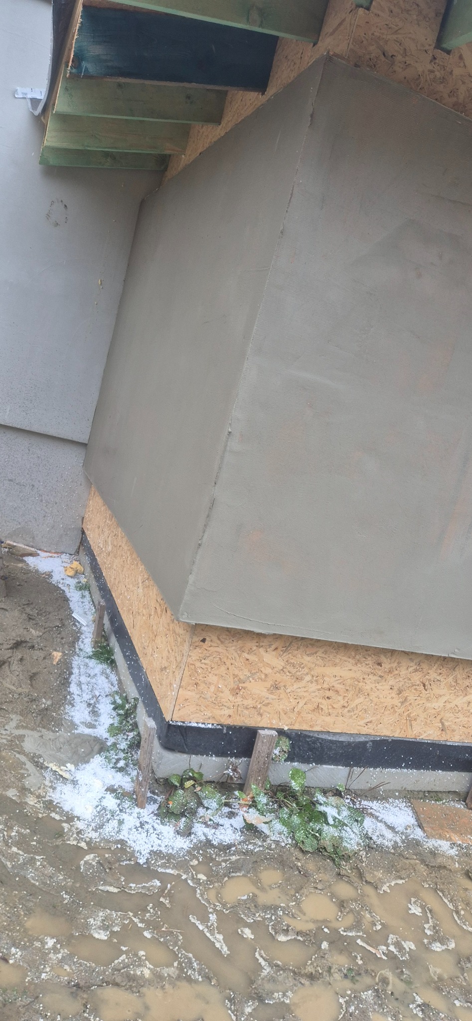 Ocieplenie fundamentu budynku z widoczną warstwą izolacji, płytą OSB i świeżo nałożonym tynkiem. Wokół błoto i resztki styropianu.