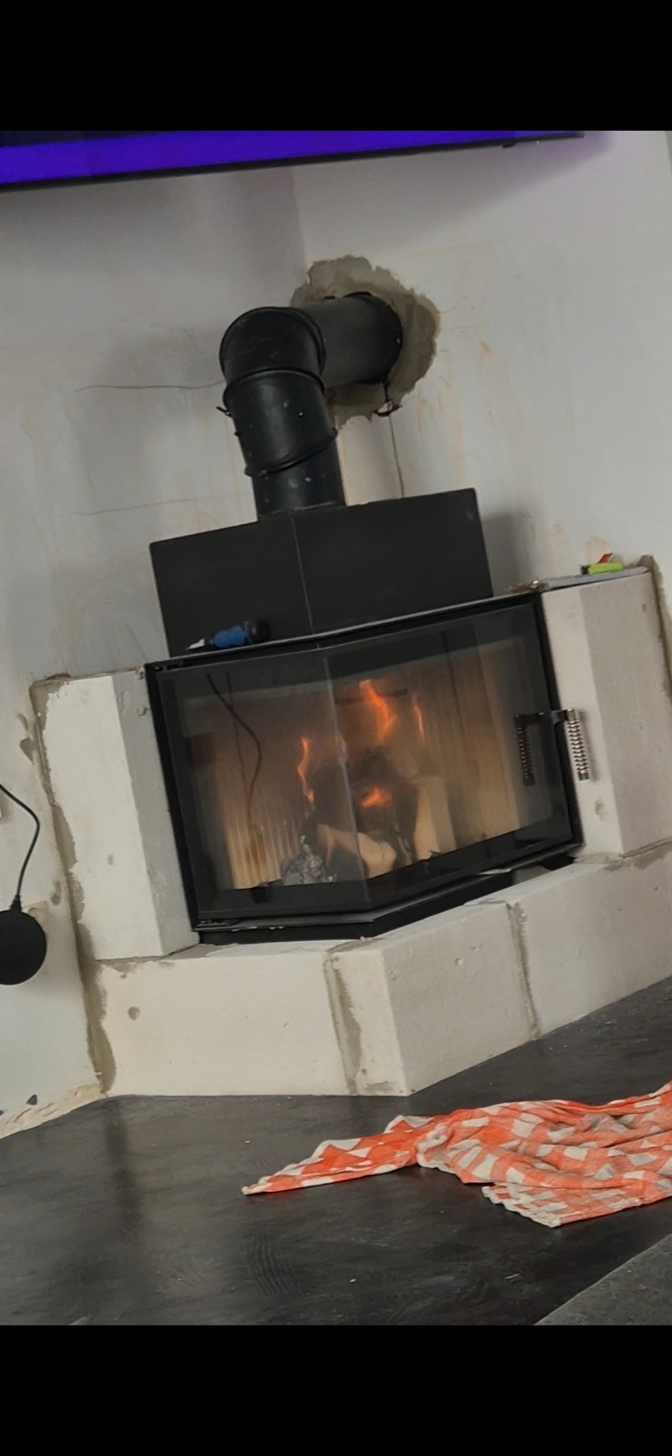 Wnętrze z kominkiem obudowanym białymi bloczkami, widoczne ślady prac wykończeniowych na ścianie i podłodze. Czarny przewód kominowy. Ogień w palenisku.