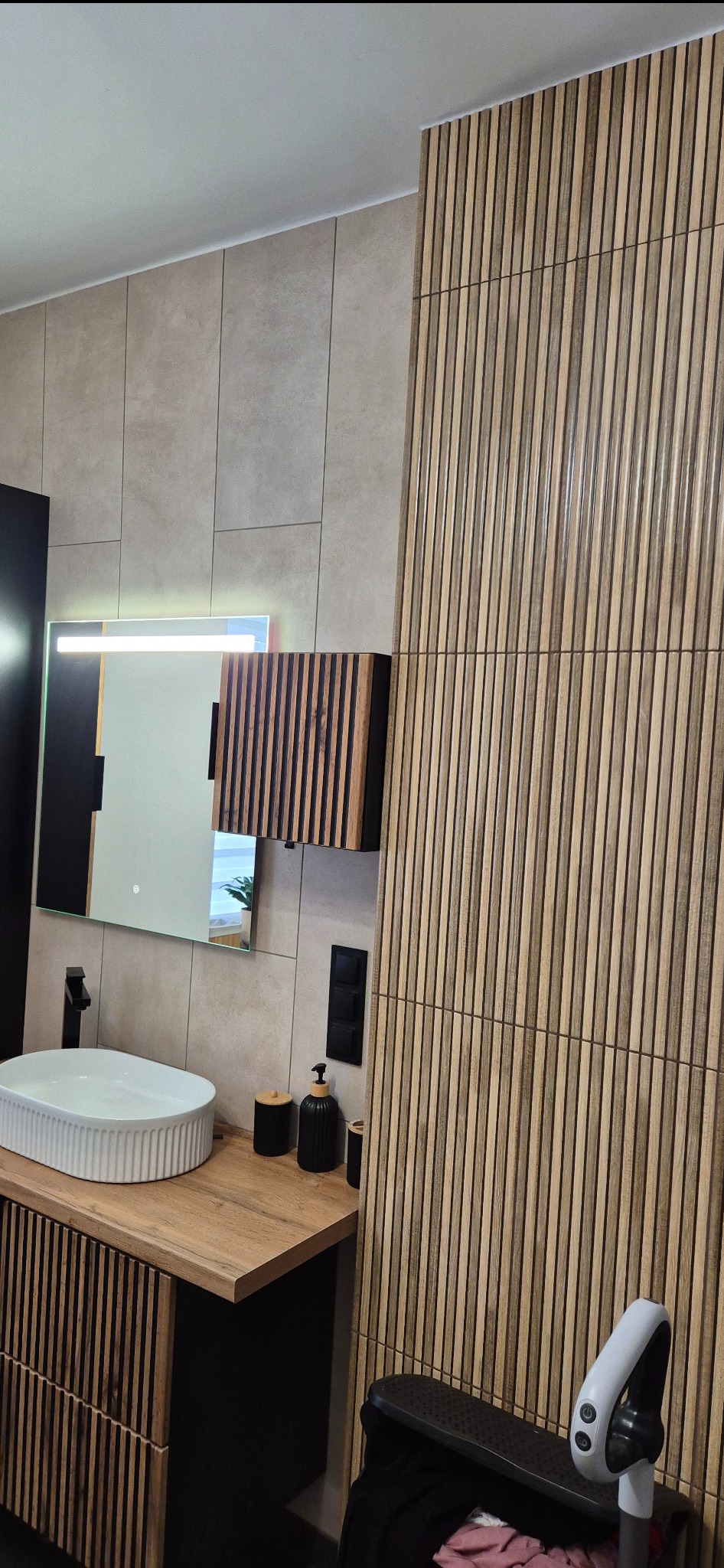 Nowoczesna łazienka z umywalką nablatową, lustrem z podświetleniem LED i ścianą wykończoną panelami dekoracyjnymi imitującymi drewno. Minimalistyczny design i ciepła kolorystyka.