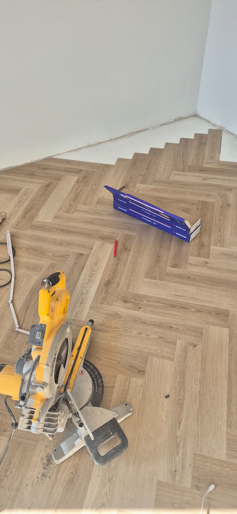 Montaż podłogi winylowej Quick-Step w jasnym odcieniu drewna, ułożonej w jodełkę, z ukośnicą DeWalt i narzędziami na pierwszym planie. Praca w rogu pomieszczenia.