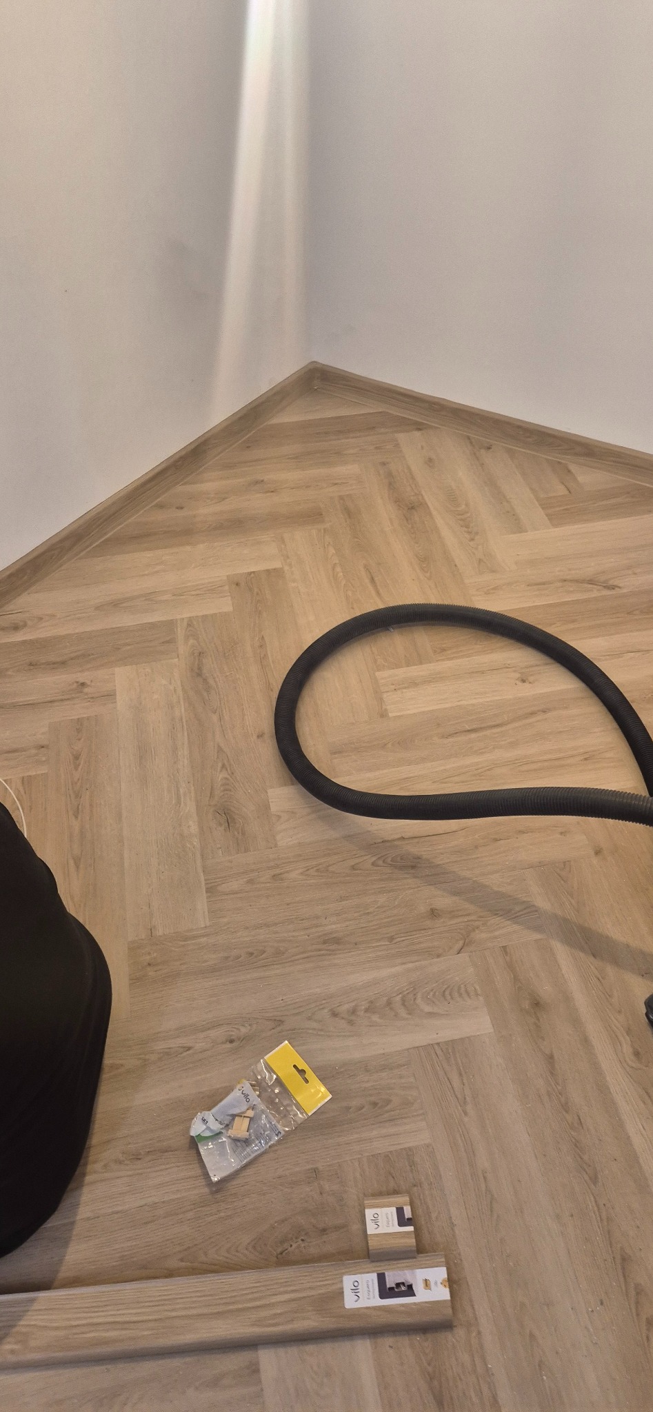 Jasna podłoga panelowa w jodełkę w narożniku pokoju, z zamontowanymi listwami przypodłogowymi Vilo i leżącym obok czarnym wężem.