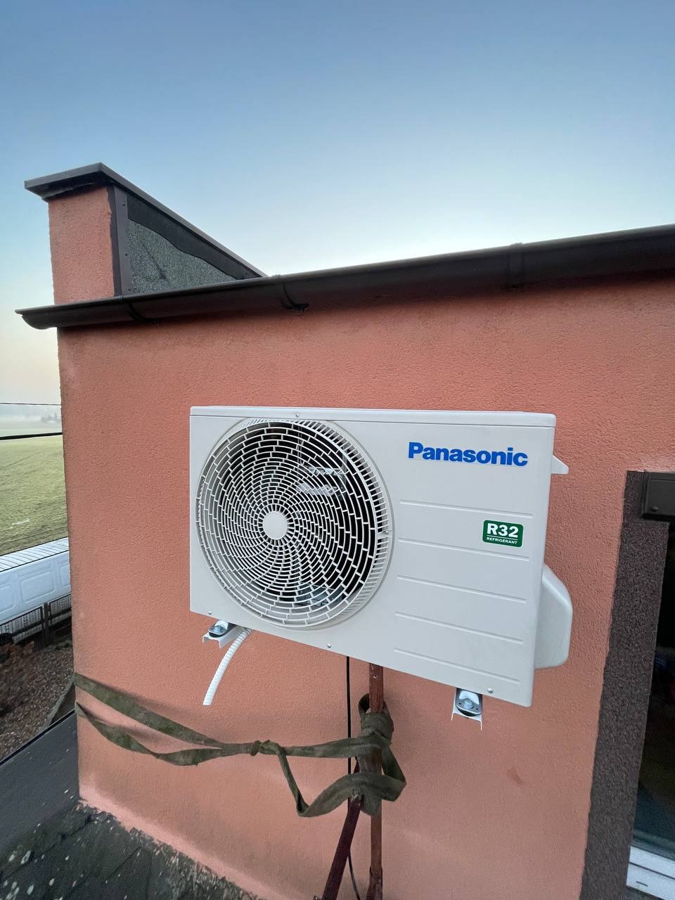 Zamontowana jednostka zewnętrzna klimatyzacji Panasonic R32 na elewacji budynku z widocznym dachem i fragmentem krajobrazu w tle. Ujęcie z dołu.