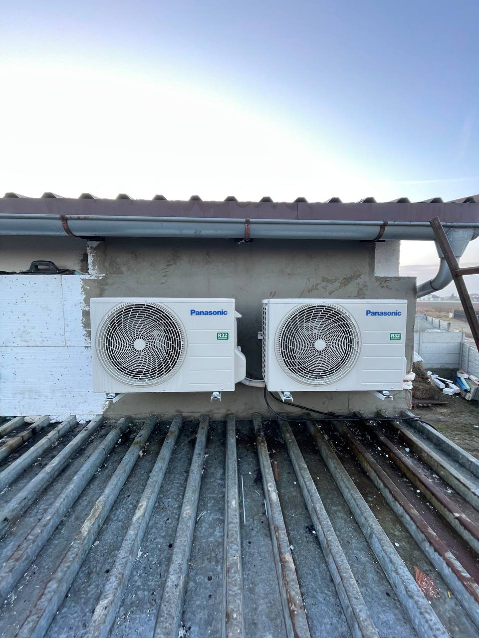 Dwa klimatyzatory Panasonic R32 zamontowane na elewacji budynku, widoczne z perspektywy dachu z metalowymi elementami konstrukcyjnymi.
