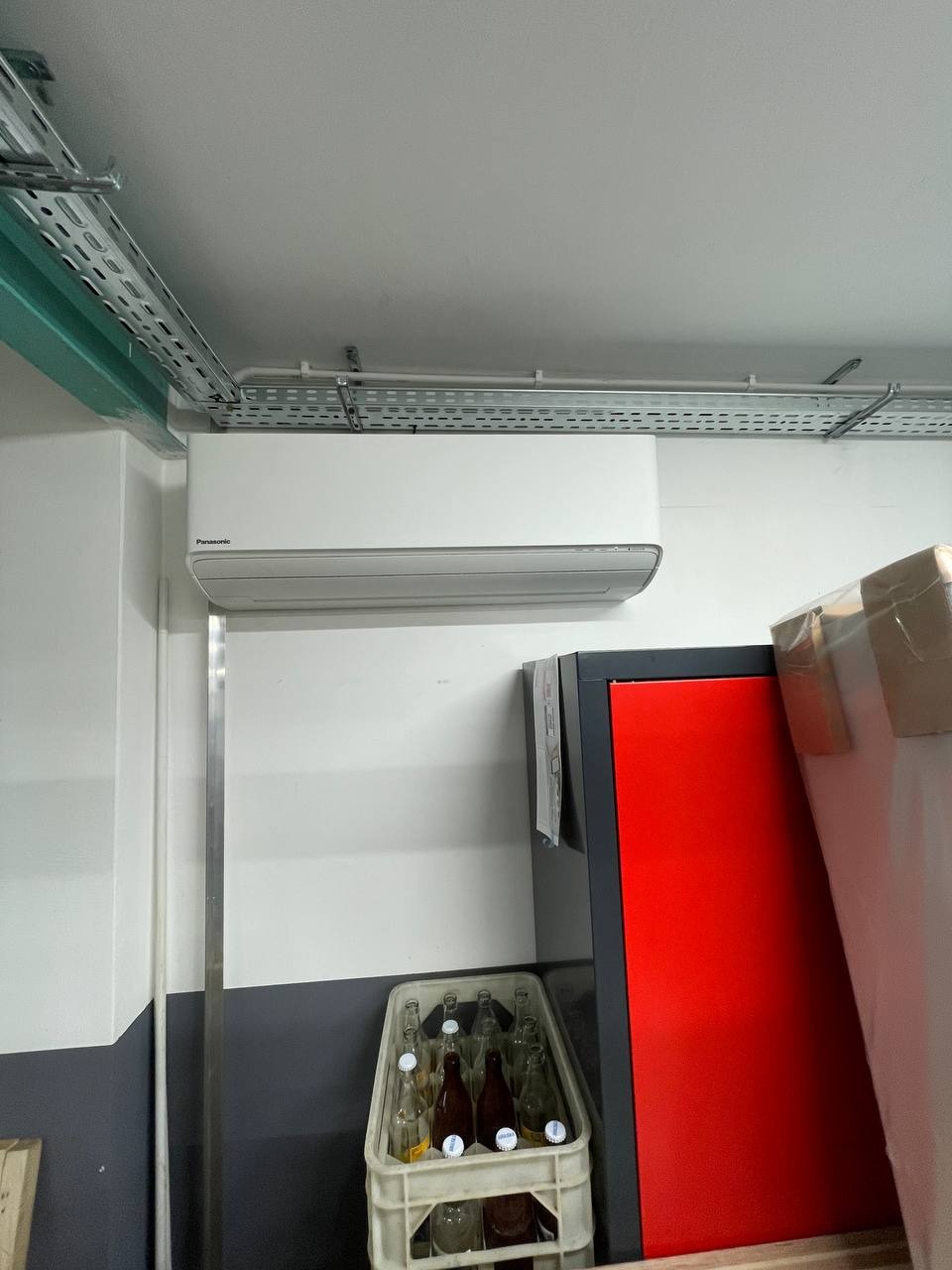 Biała klimatyzacja Panasonic zamontowana na ścianie, widoczne kable w korytkach, obok czerwona lodówka i skrzynka z butelkami. Ujęcie z dołu.