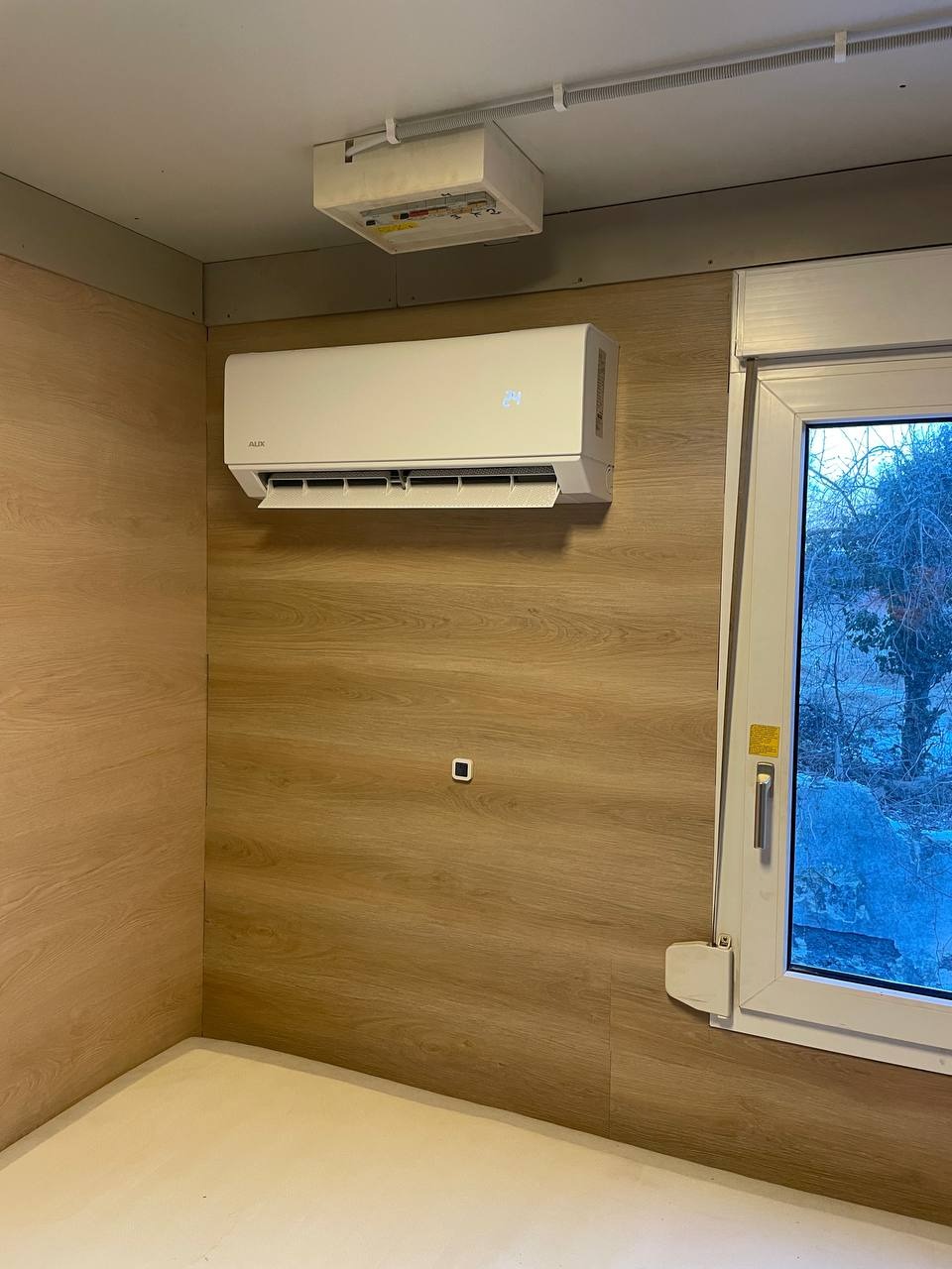 Biały klimatyzator AUX zamontowany na ścianie z panelami drewnopodobnymi w jasnym pomieszczeniu z oknem. Widoczna instalacja elektryczna pod sufitem.