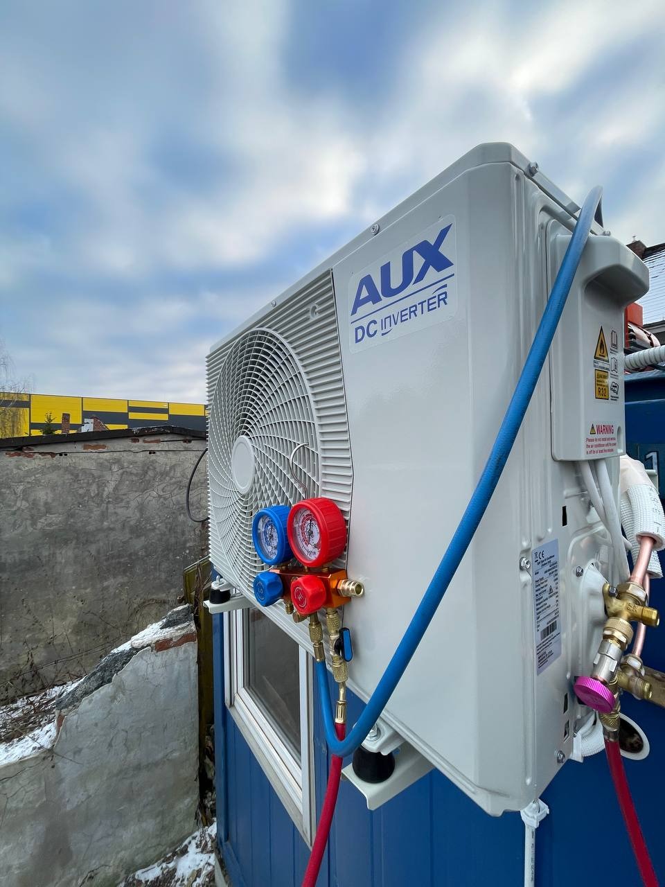 Biały klimatyzator AUX DC Inverter zamontowany na zewnątrz budynku, z podłączonymi przewodami i manometrami, na tle pochmurnego nieba.