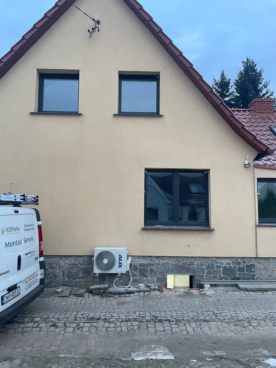 Montaż klimatyzacji zewnętrznej AUX przy beżowej elewacji domu z czarnymi oknami. Widoczny fragment firmowego busa z logo KliMate i numerem telefonu.