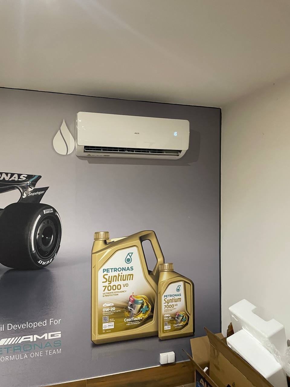 Biała klimatyzacja AUX na szarej ścianie z grafiką bolidu Formuły 1 i oleju Petronas Syntium 7000. Widoczny wyświetlacz temperatury.
