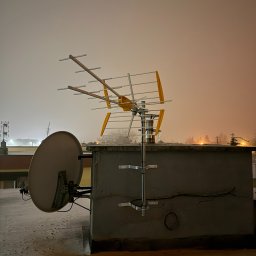 NETHOME MARCIN KOSTRZEWSKI - Antena DVB-T2 i satelitarna na dachu budynku w zimowej scenerii. Widoczne elementy montażowe i przewody. Nocne oświetlenie w tle.