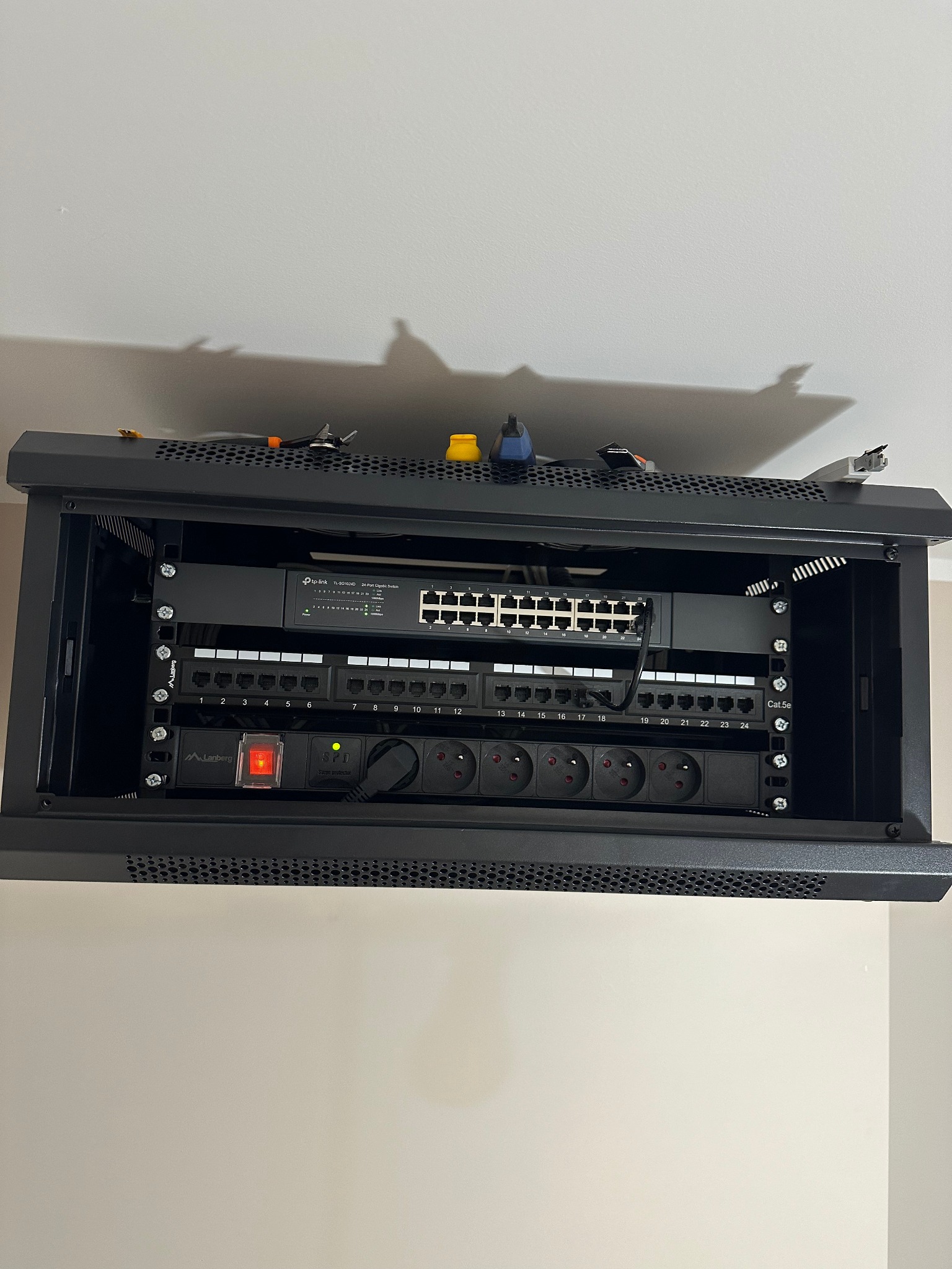 Zamknięta szafa rack z zamontowanym switchem TP-Link, panelem krosowniczym i listwą zasilającą Lanberg, zainstalowana na ścianie. Widoczne kable i akcesoria.
