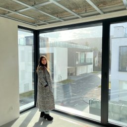 STUDIO K Karolina Leszczak - Wnętrze w stanie deweloperskim z widoczną konstrukcją sufitu podwieszanego i dużym oknem narożnym. Kobieta w płaszczu stoi w jasnym pomieszczeniu.