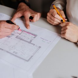 STUDIO K Karolina Leszczak - Osoby pracują nad planem mieszkania, używając markerów i ołówka do zaznaczania szczegółów. Widoczne dłonie, fragment planu z naniesionymi notatkami i elementami wyposażenia.