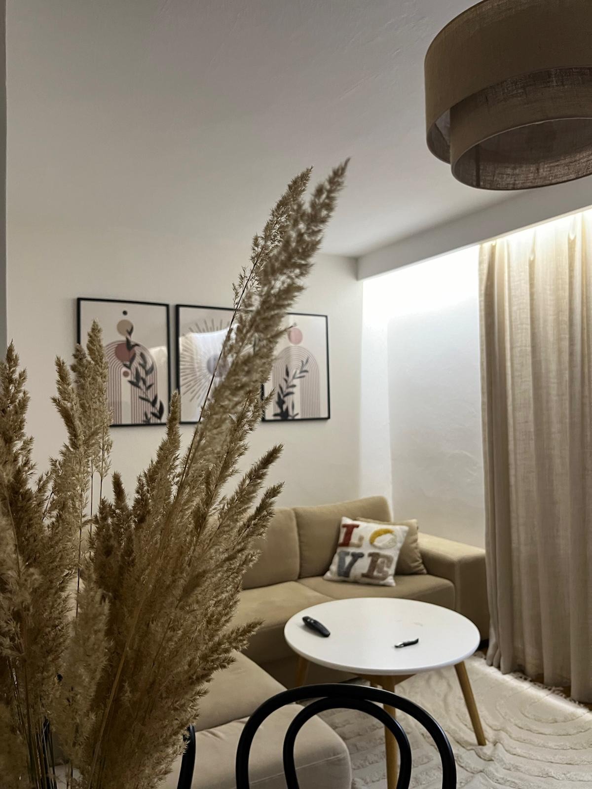 Jasne wnętrze salonu z beżową sofą, okrągłym stolikiem i dekoracyjnymi obrazami na ścianie. Wazon z suszonymi trawami na pierwszym planie dodaje przytulności.