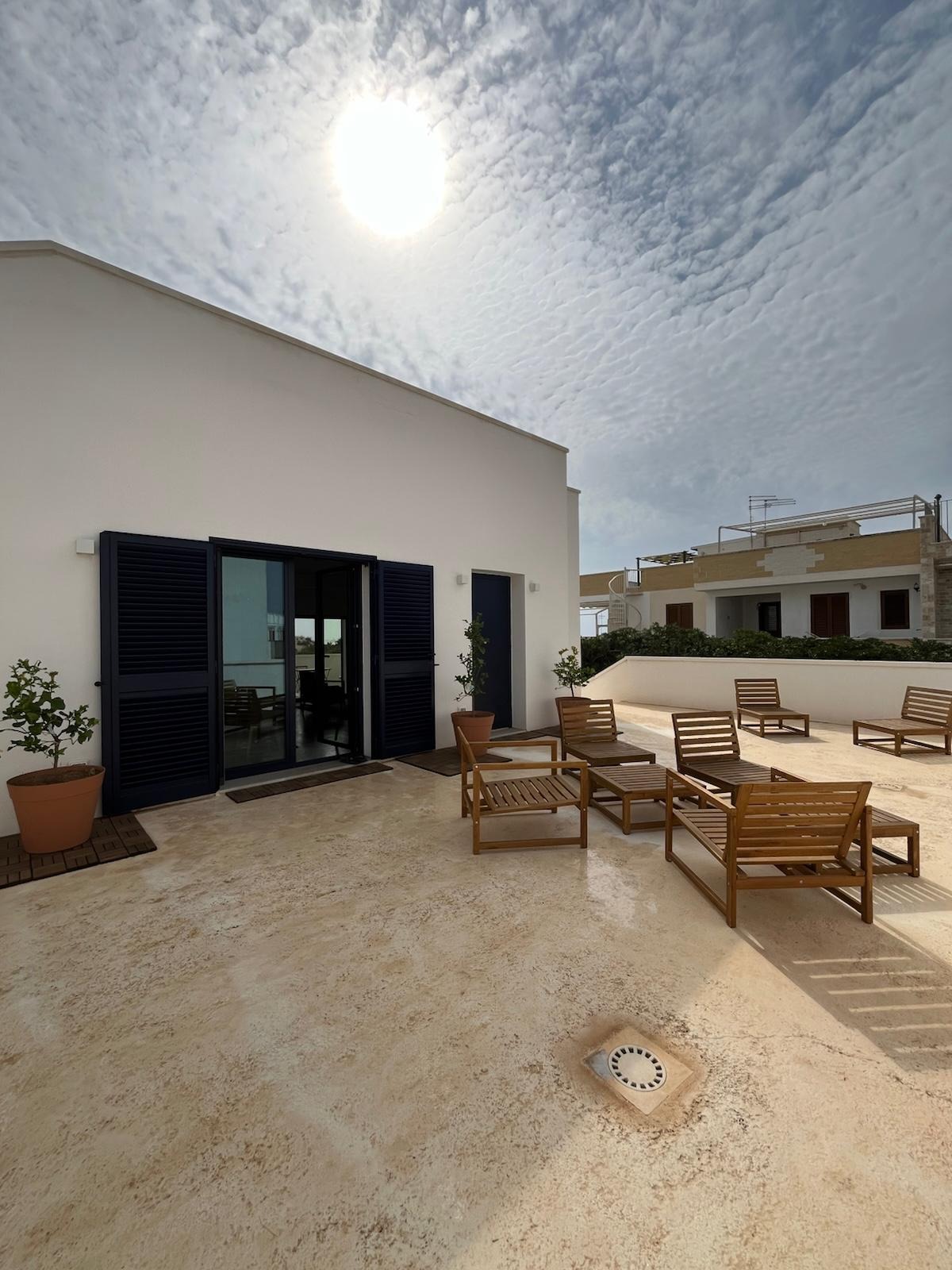 Biały budynek z ciemnymi okiennicami i drewnianymi meblami ogrodowymi na tarasie w słoneczny dzień. Minimalistyczny design z roślinami w donicach.