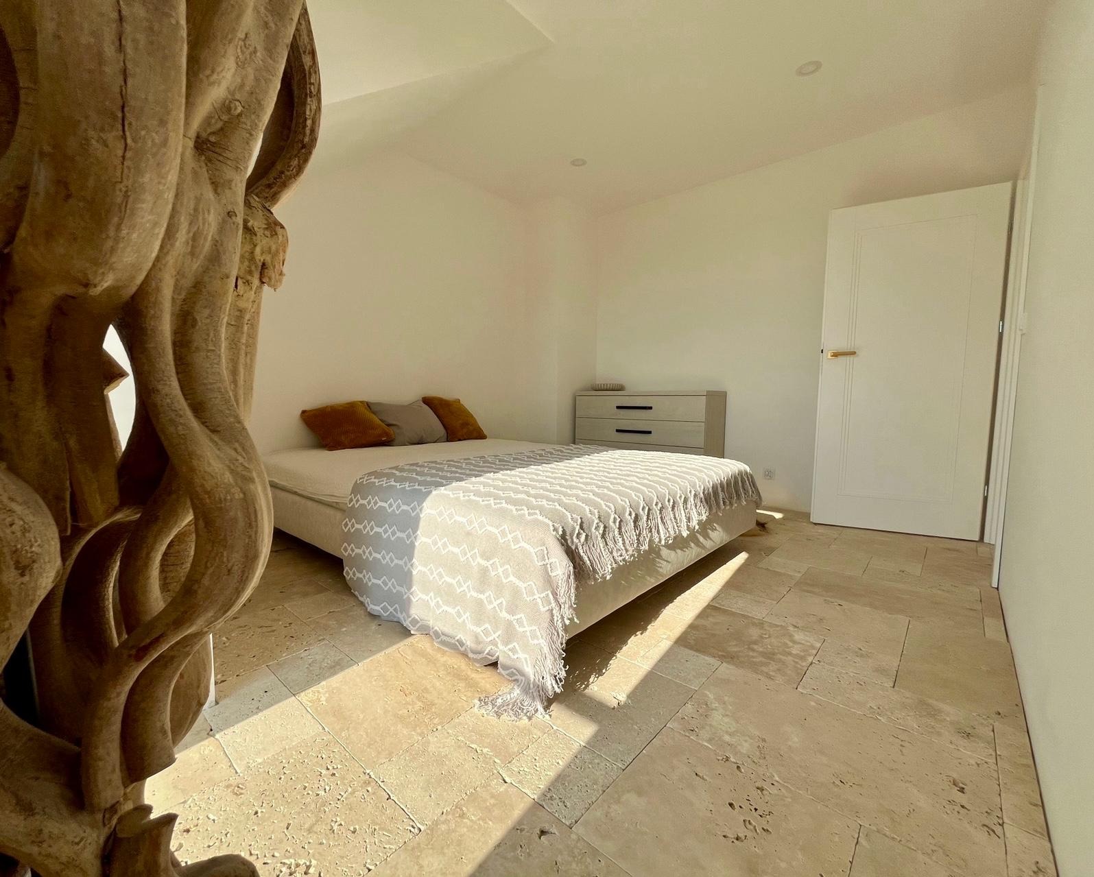 Jasna sypialnia z beżową podłogą z kamienia, drewnianą rzeźbą i łóżkiem z narzutą w geometryczne wzory. Widoczne promienie słoneczne.