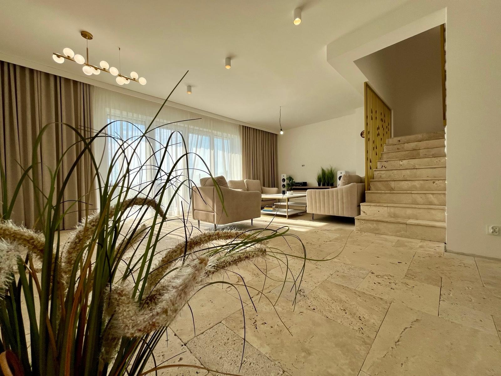 Jasne wnętrze salonu z beżowymi sofami, marmurową podłogą i schodami. Dekoracyjna trawa w wazonie na pierwszym planie, złote akcenty oświetlenia i balustrady.