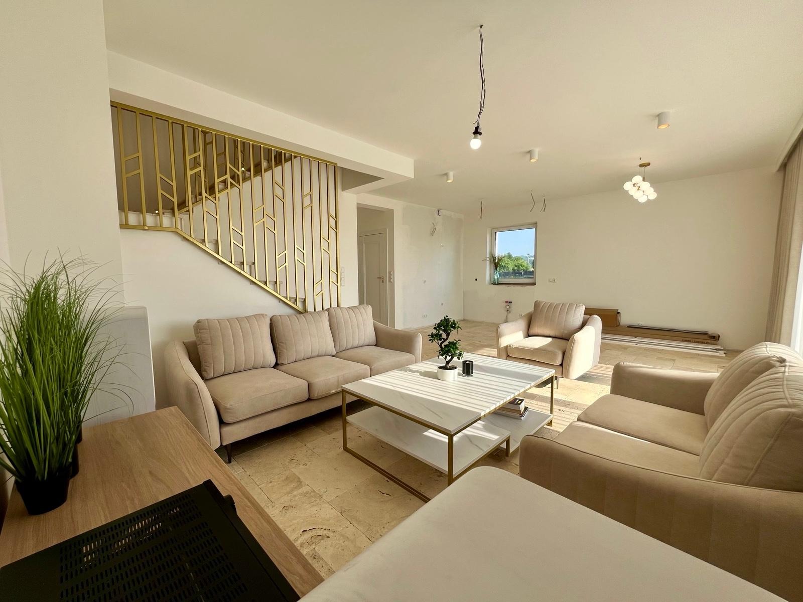 Jasne wnętrze salonu z beżowymi sofami, marmurowym stolikiem i złotą balustradą schodów. Podłoga wyłożona płytkami imitującymi kamień. Widoczne niedokończone elementy.