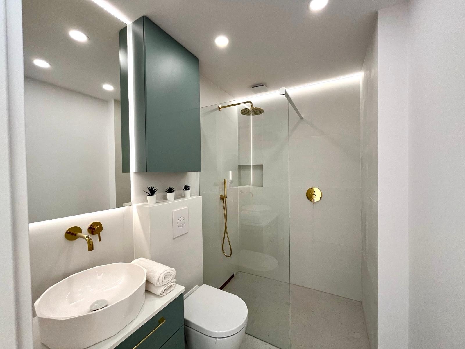 Nowoczesna łazienka z kabiną prysznicową typu walk-in, umywalką nablatową i szafką w odcieniach zieleni. Złote akcenty armatury i oświetlenie LED podkreślają elegancki design.
