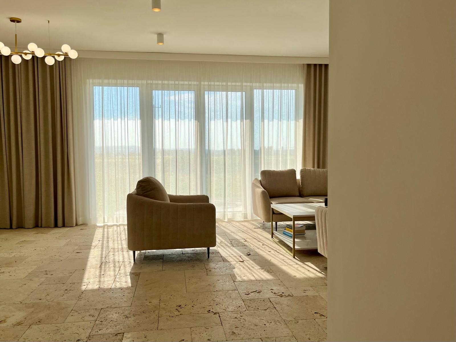 Jasne wnętrze salonu z trawertynową podłogą, beżowymi zasłonami i meblami w odcieniach brązu. Promienie słońca wpadają przez okna.