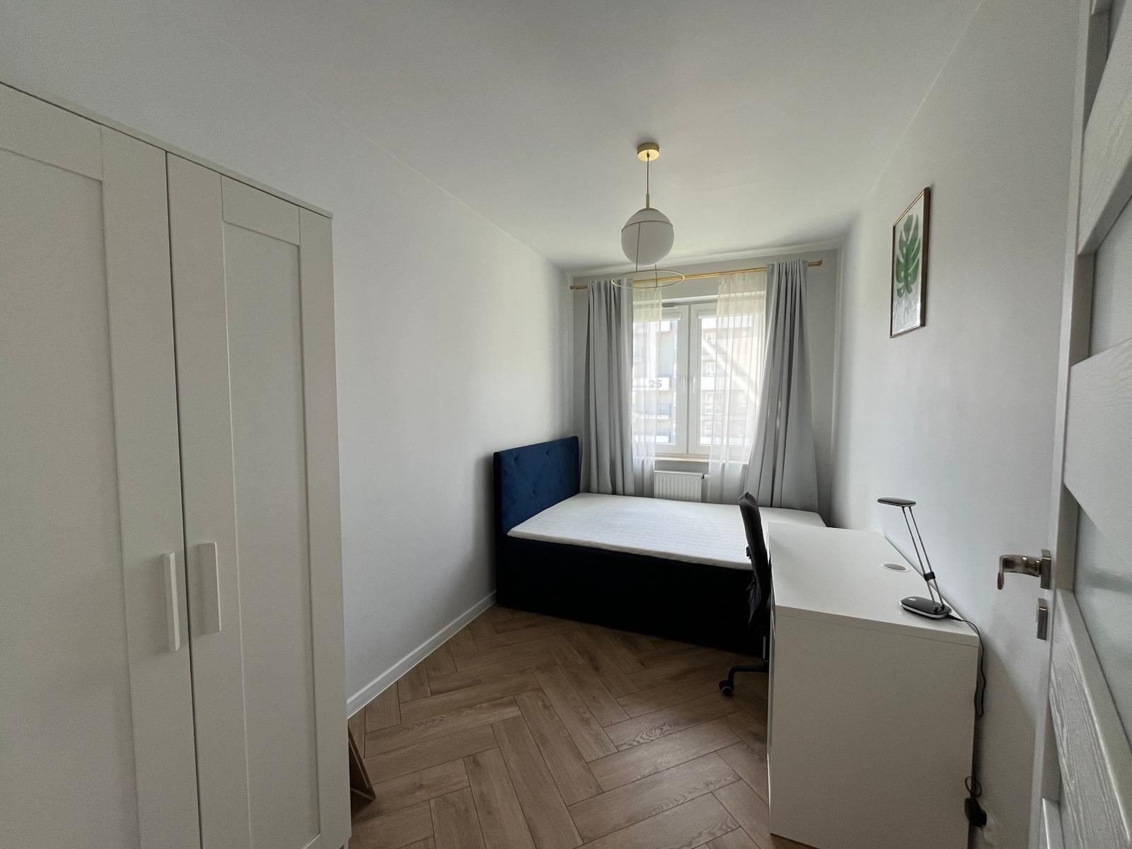 Pokój po remoncie: parkiet w jodełkę, białe ściany, szafa, biurko i łóżko z niebieskim zagłówkiem. Minimalistyczny wystrój, okno z szarymi zasłonami.