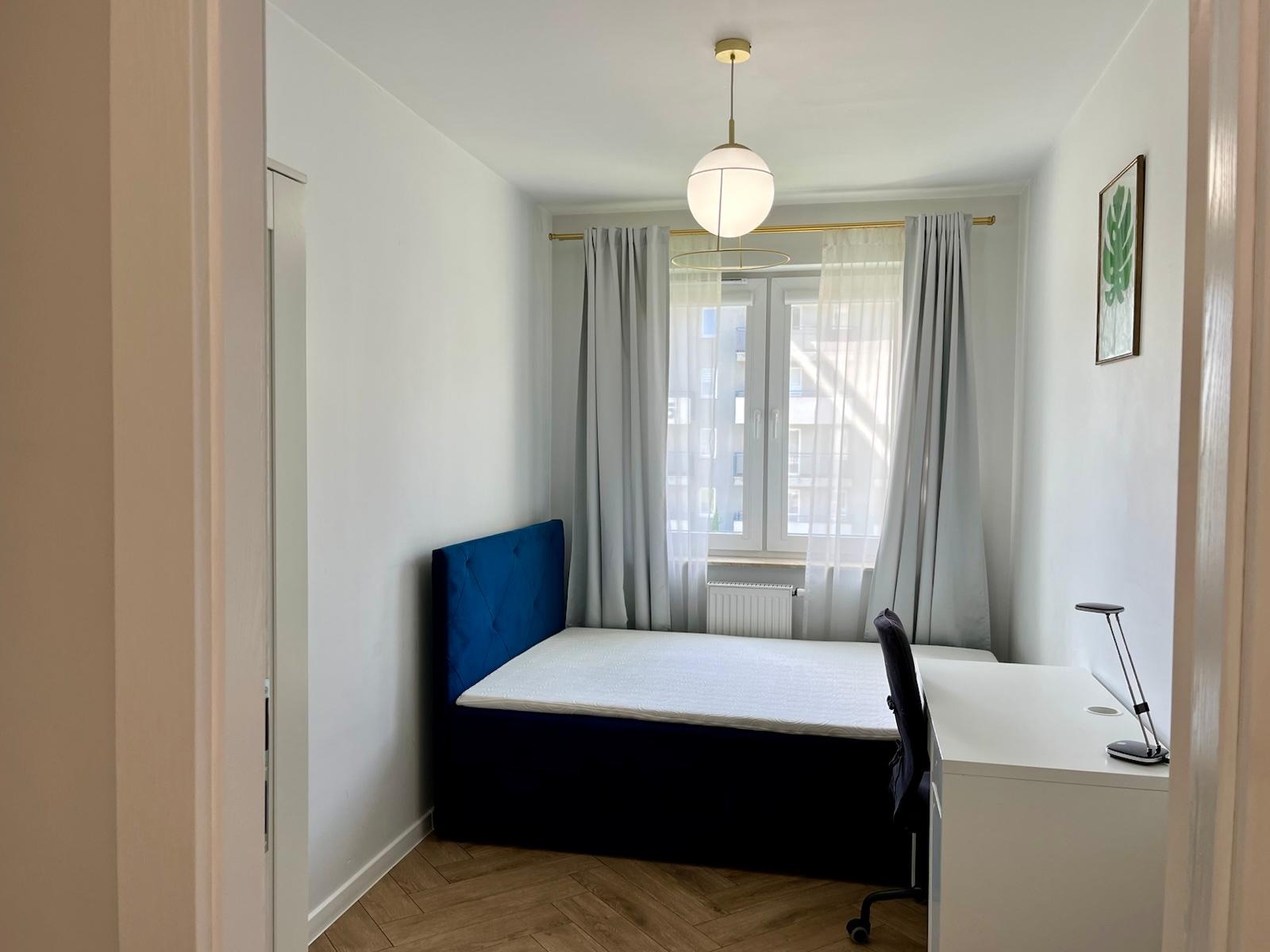 Jasny pokój z łóżkiem z niebieskim wezgłowiem, biurkiem i parkietem ułożonym w jodełkę. Okno z szarymi zasłonami i nowoczesna lampa wisząca.