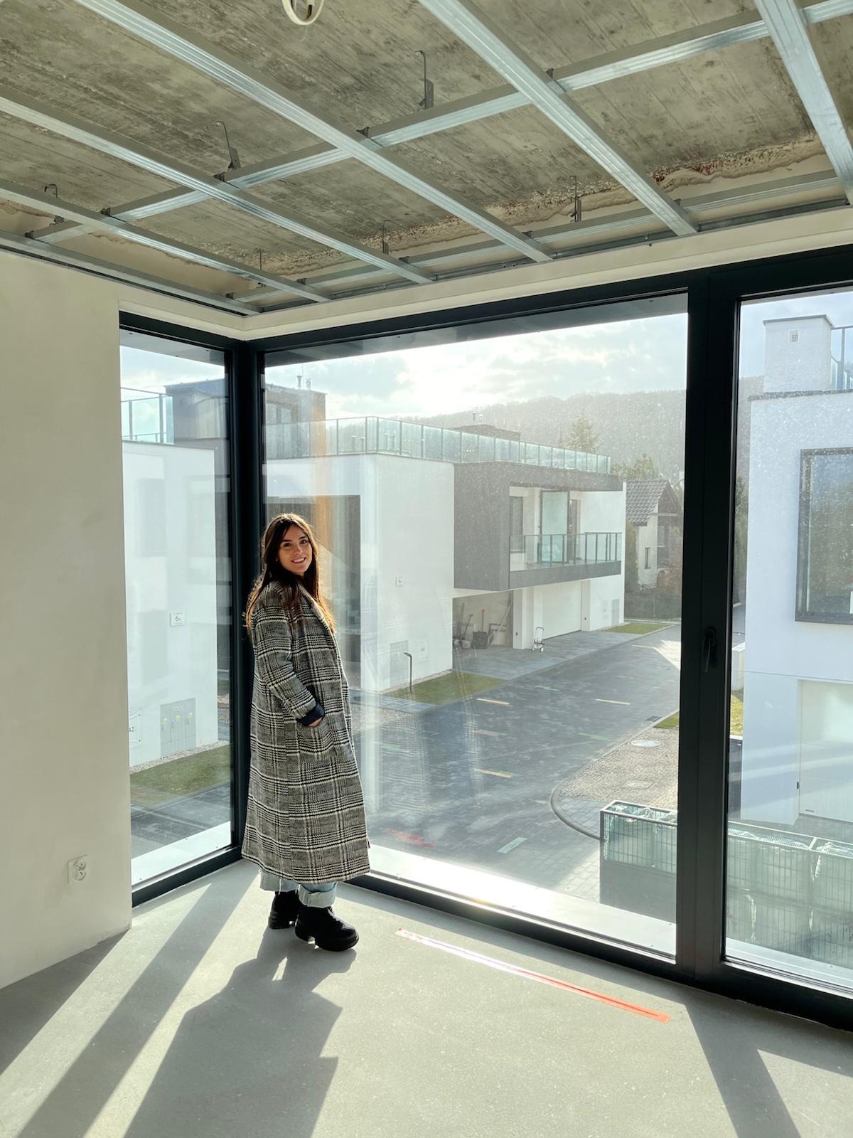 Wnętrze w stanie deweloperskim z widoczną konstrukcją sufitu podwieszanego i dużym oknem narożnym. Kobieta w płaszczu stoi w jasnym pomieszczeniu.