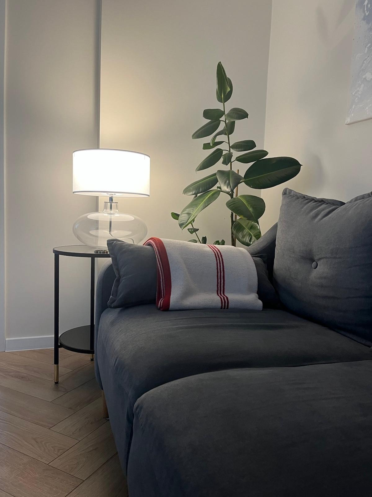 Stylowy salon z szarą sofą, stolikiem z lampą i rośliną doniczkową. Parkiet ułożony w jodełkę. Przytulne wnętrze z dbałością o detale.