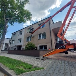Klimler Leraczyk Marcin - Praca na wysokości: pomarańczowy podnośnik koszowy przy budynku, osoba w koszu przy klimatyzatorze. Widoczne drzewo i chodnik z kostki.