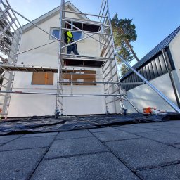 Klimler Leraczyk Marcin - Pracownik na rusztowaniu maluje elewację domu. Widoczne zabezpieczone okna, narzędzia i wiadro z farbą. Ujęcie z dołu, podkreślające wysokość rusztowania.