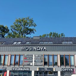 Klimler Leraczyk Marcin - Budynek z panelami słonecznymi na dachu, widok z dołu w słoneczny dzień. Na fasadzie szyldy firm i okna. U góry drzewa i błękitne niebo.