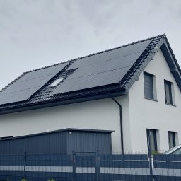 Biały dom jednorodzinny z ciemną dachówką i panelami słonecznymi na dachu, widok z poziomu gruntu, pochmurne niebo w tle.