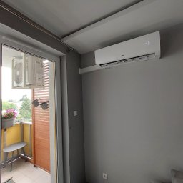 Biały klimatyzator CH na szarej ścianie w pokoju, widok na balkon z kwiatami i krzesłem przez otwarte drzwi balkonowe. Nowoczesny design, wnętrze mieszkania.