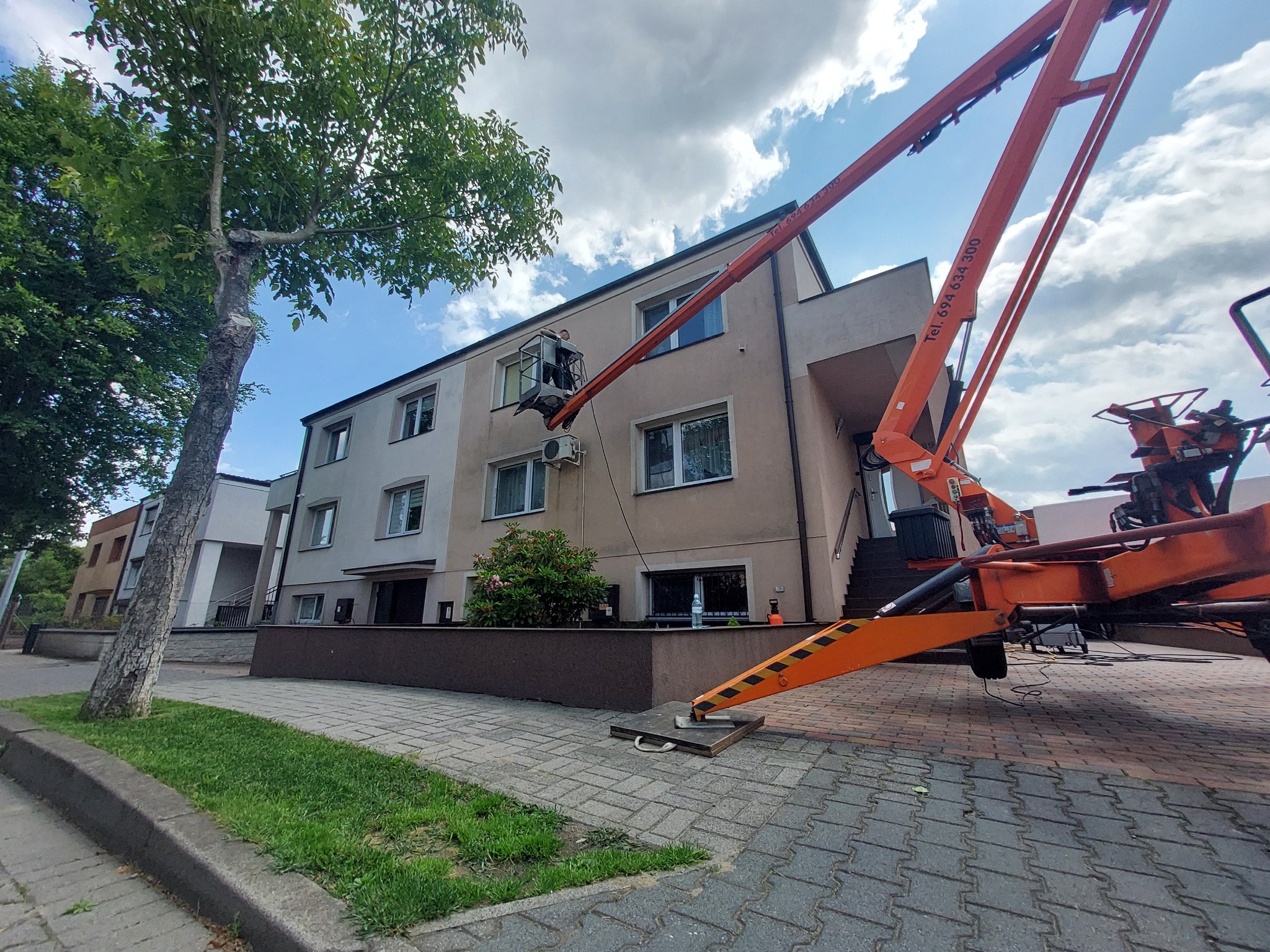Praca na wysokości: pomarańczowy podnośnik koszowy przy budynku, osoba w koszu przy klimatyzatorze. Widoczne drzewo i chodnik z kostki.