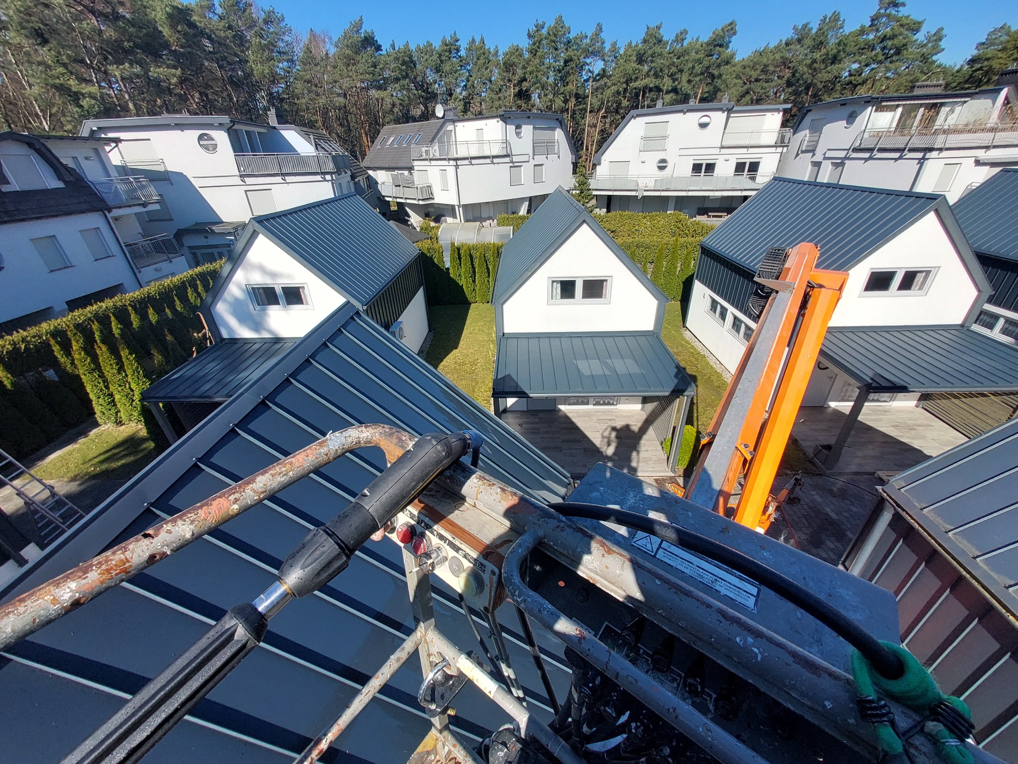 Widok z podnośnika koszowego na osiedle domów jednorodzinnych z ciemnymi dachami w słoneczny dzień. W kadrze fragment urządzenia podnośnika.