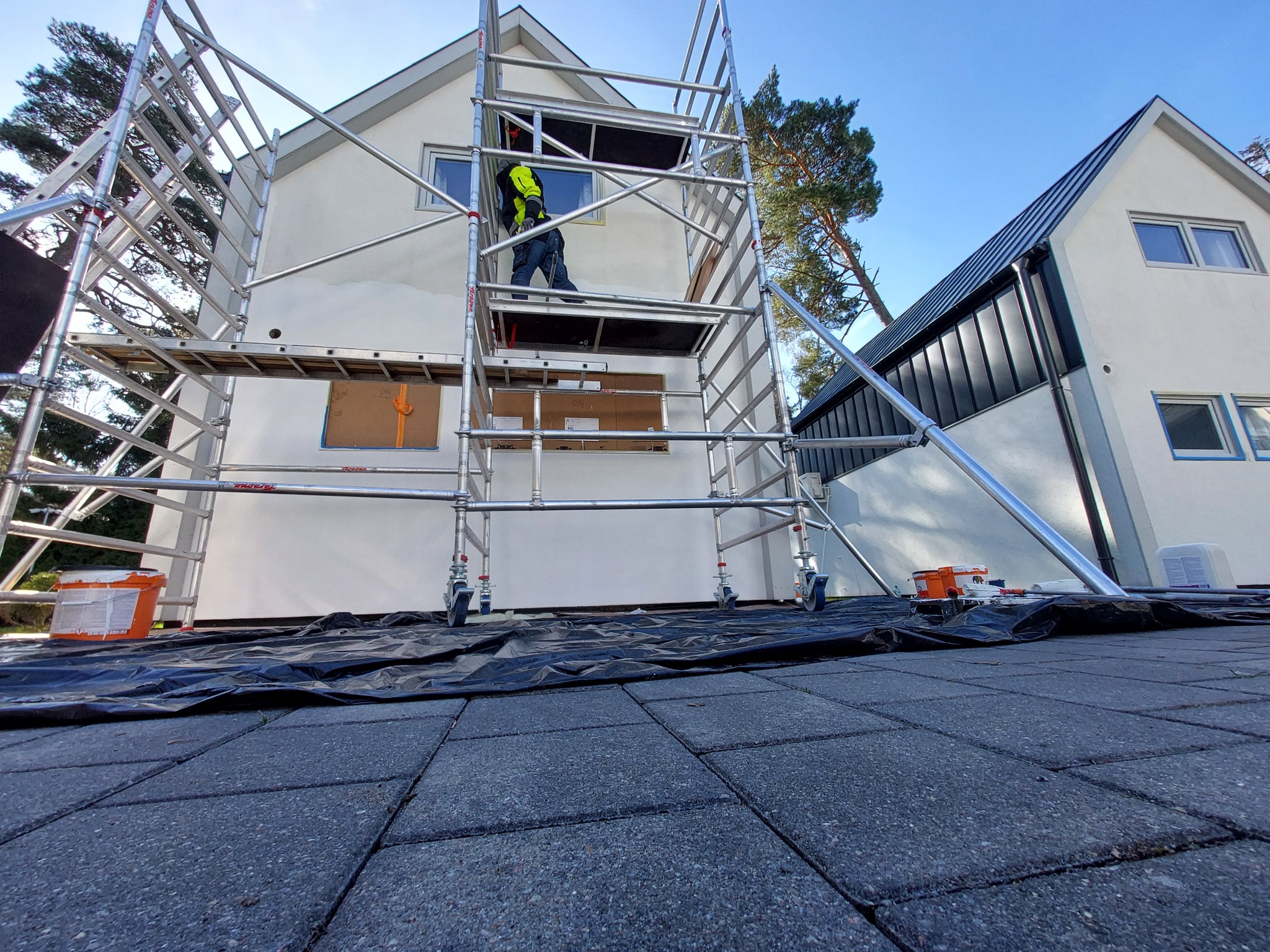 Pracownik na rusztowaniu maluje elewację domu. Widoczne zabezpieczone okna, narzędzia i wiadro z farbą. Ujęcie z dołu, podkreślające wysokość rusztowania.