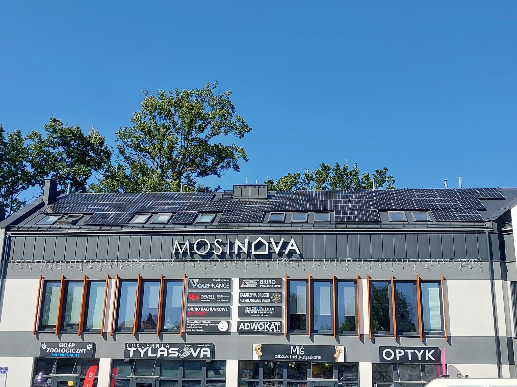 Budynek z panelami słonecznymi na dachu, widok z dołu w słoneczny dzień. Na fasadzie szyldy firm i okna. U góry drzewa i błękitne niebo.