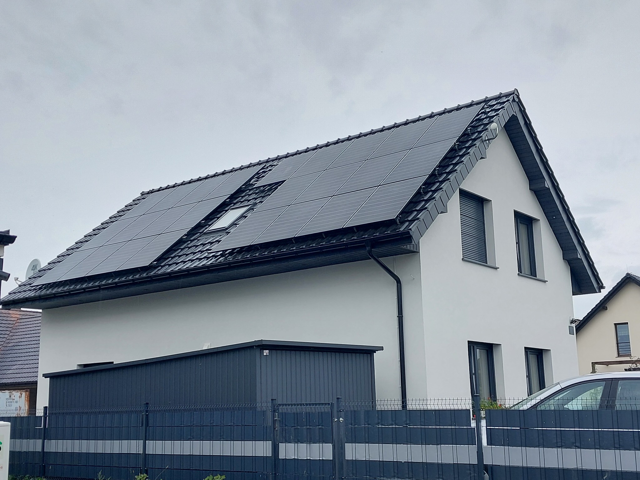 Biały dom jednorodzinny z ciemną dachówką i panelami słonecznymi na dachu, widok z poziomu gruntu, pochmurne niebo w tle.