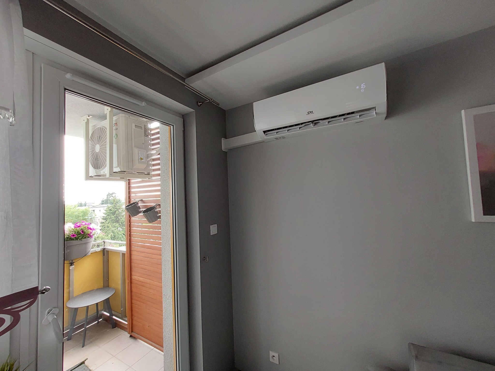 Biały klimatyzator CH na szarej ścianie w pokoju, widok na balkon z kwiatami i krzesłem przez otwarte drzwi balkonowe. Nowoczesny design, wnętrze mieszkania.